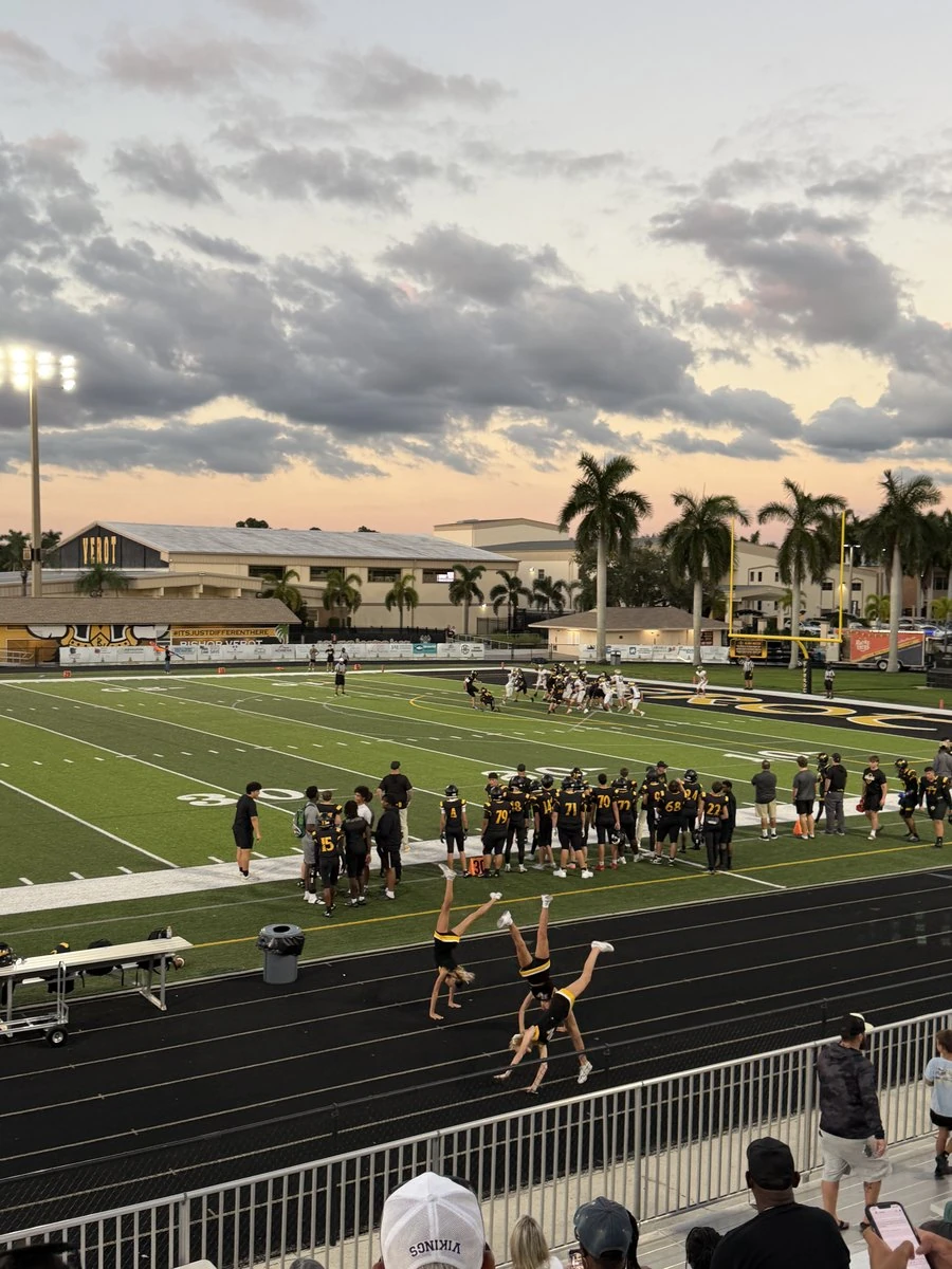 A home football game at dusk, photographed from the stands, with the team on the sideline and cheerleaders on the track